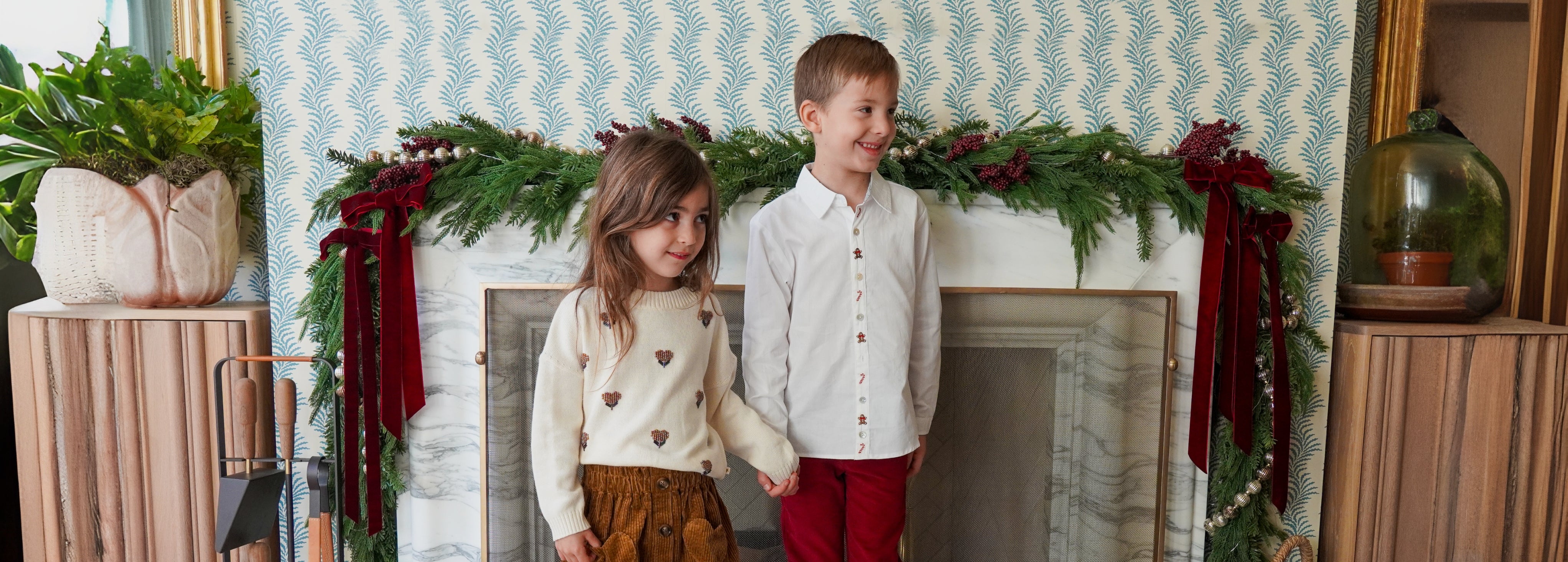 Young girl wearing a cream sweater with embroidered floral details standing beside a boy in a white button-down shirt with festive embroidery, in front of a fireplace decorated with evergreen garlands and burgundy velvet bows.