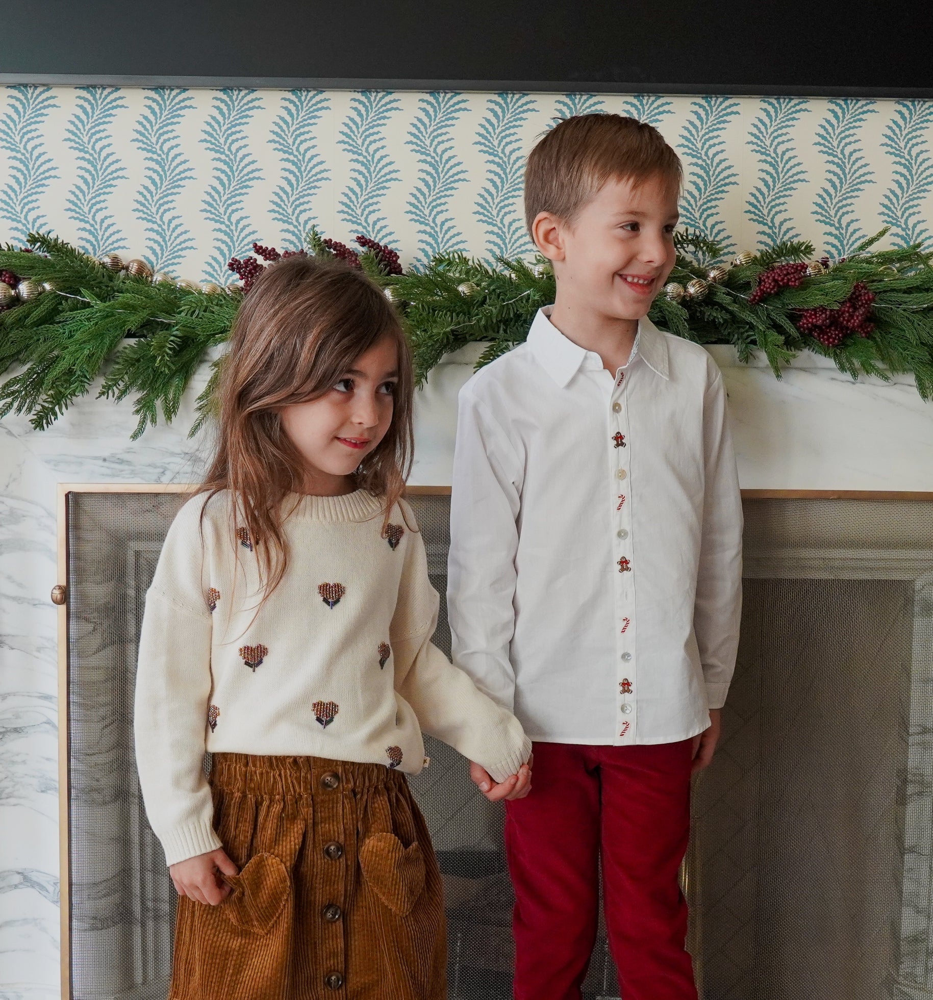 Young girl wearing a cream sweater with embroidered floral details standing beside a boy in a white button-down shirt with festive embroidery, in front of a fireplace decorated with evergreen garlands and burgundy velvet bows.
