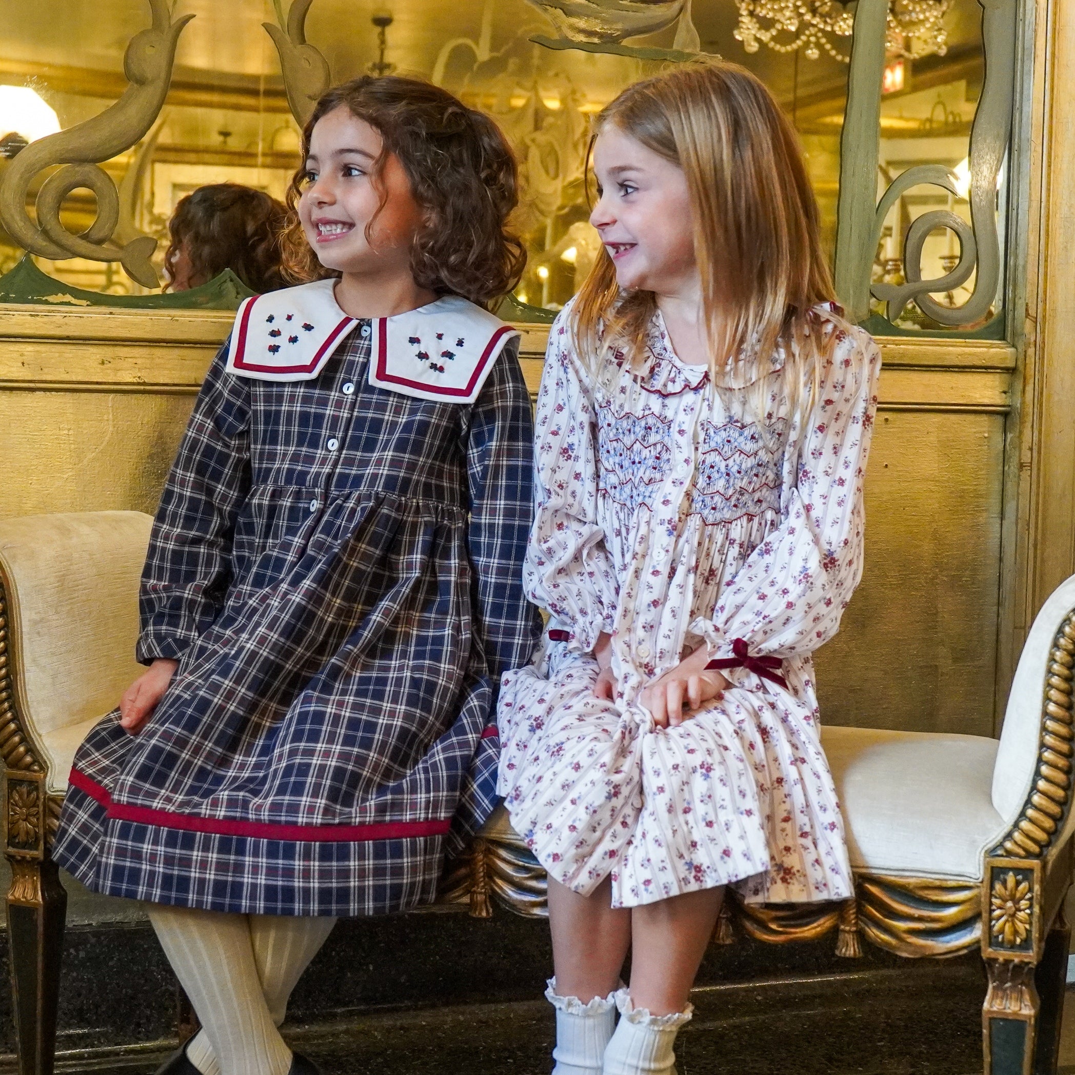 Two smiling young girls sitting together on an elegant gold-trimmed bench, wearing festive navy plaid and white floral dresses with embroidered collars and burgundy accents.