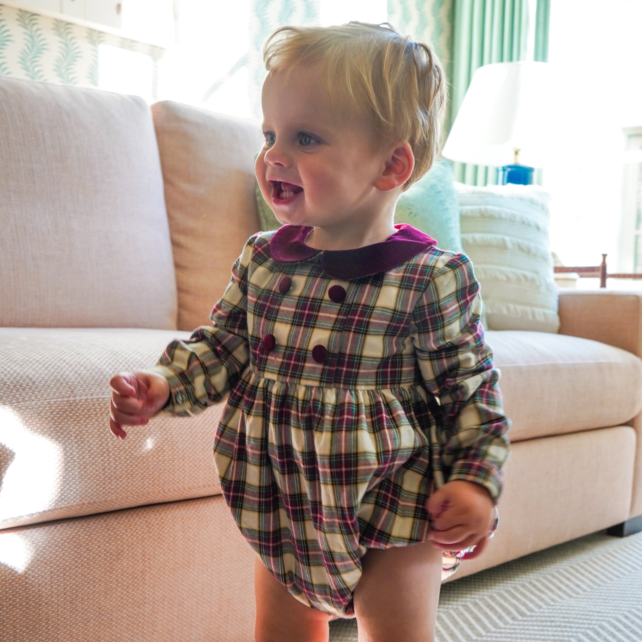Smiling toddler standing beside a cream sofa wearing a festive plaid bubble romper with long sleeves, burgundy velvet collar, and matching buttons.