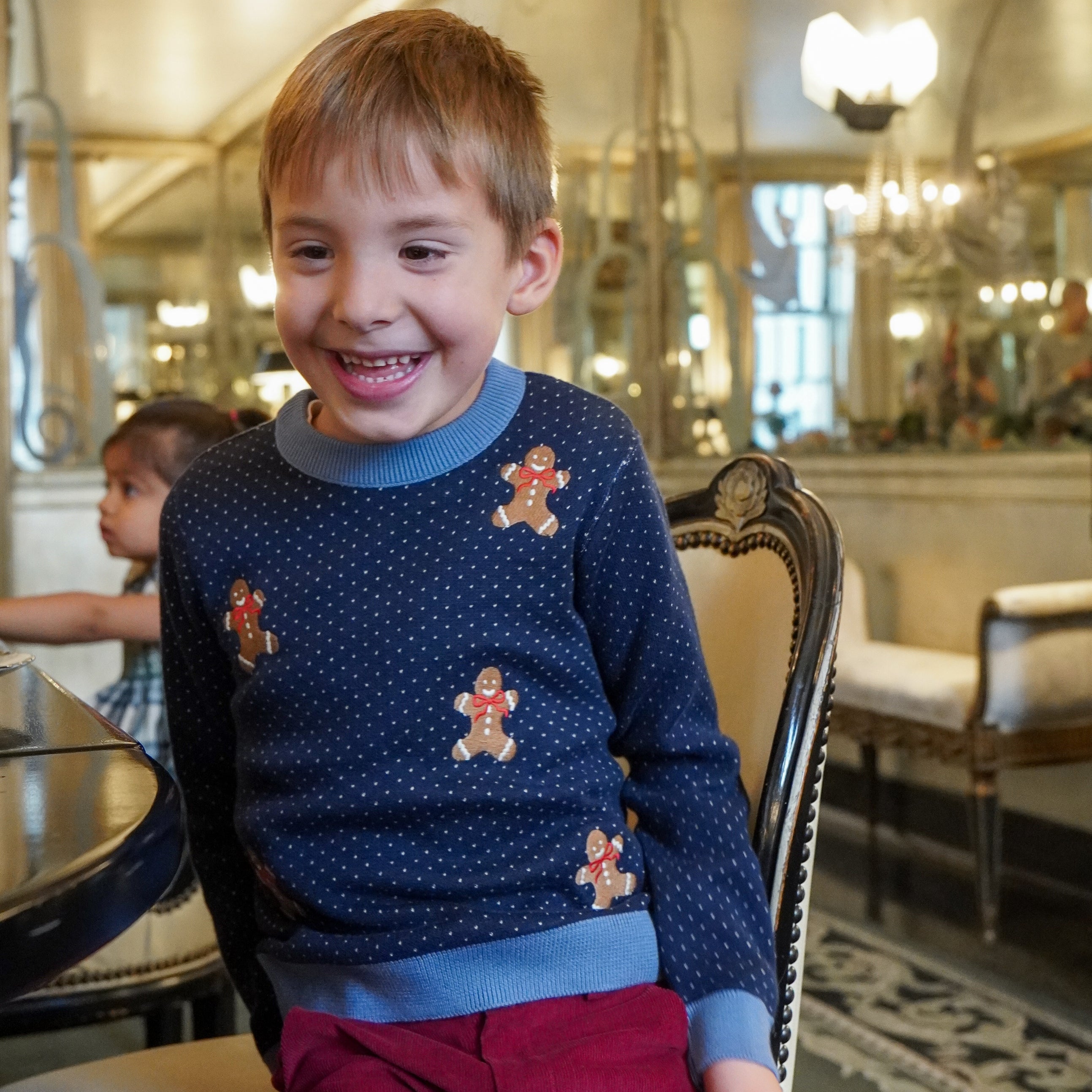 Smiling young boy wearing a navy blue sweater with gingerbread man embroidery, light blue trim, and red pants.