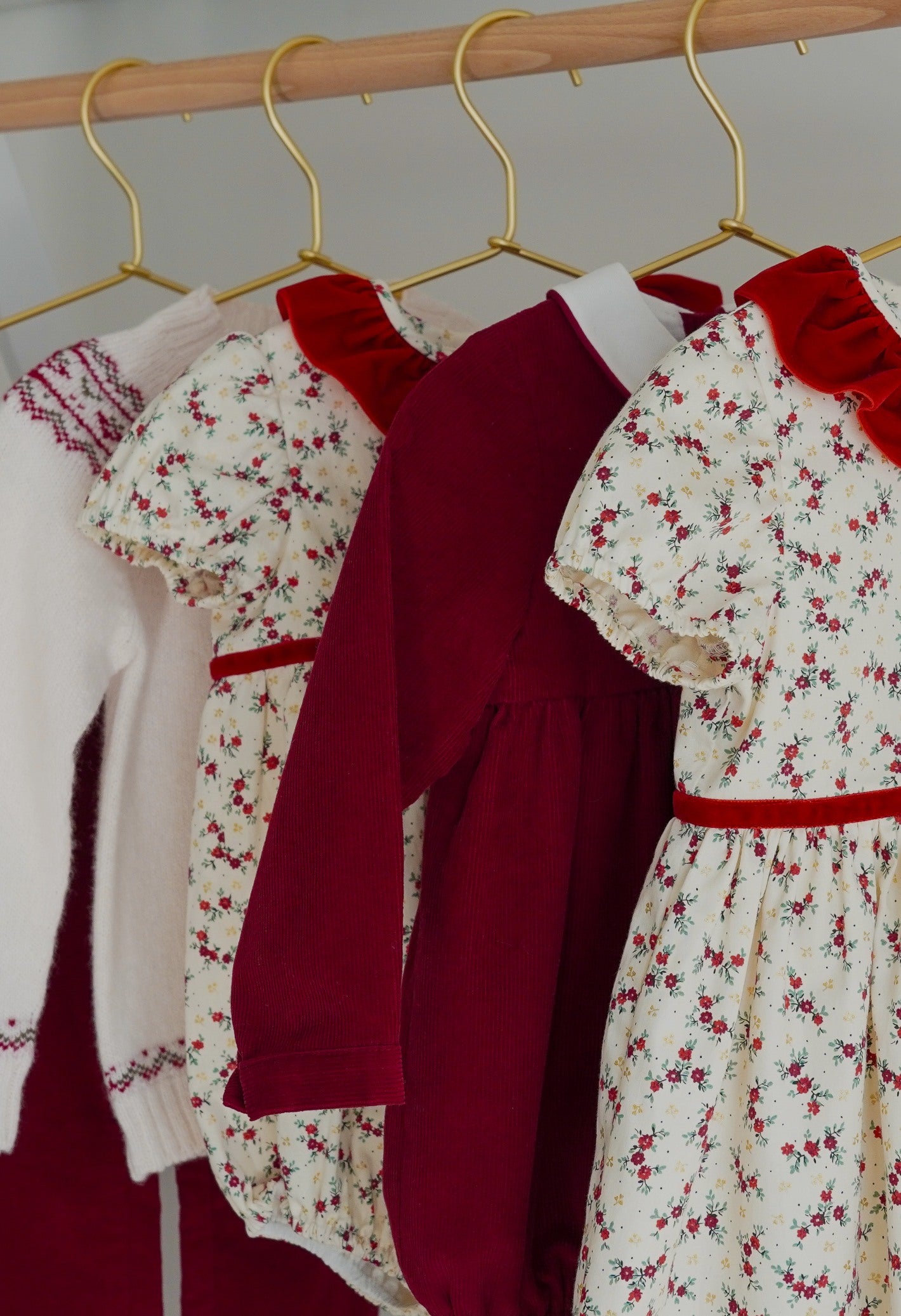 A row of children's clothing hanging on gold hangers from a wooden rod, including cream floral dresses with red collars and belts, a solid burgundy long-sleeve top, and a white knit sweater with red trim.