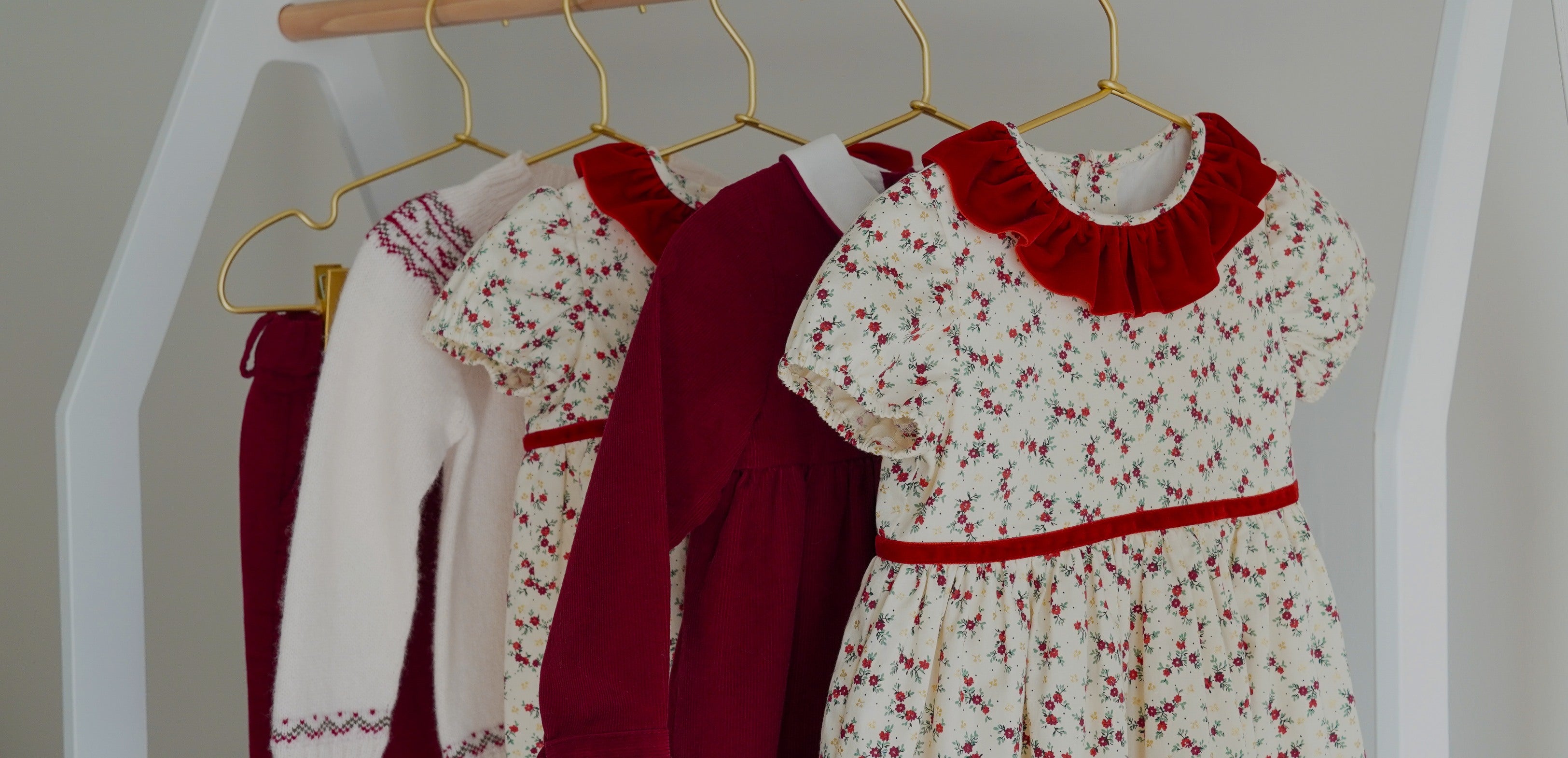 A set of children's clothing hanging on a wooden hanger rack, featuring a cream dress with a red floral print and red ruffle collar, a dark red long-sleeve top, a cream floral dress with a red collar, and dark red trousers.