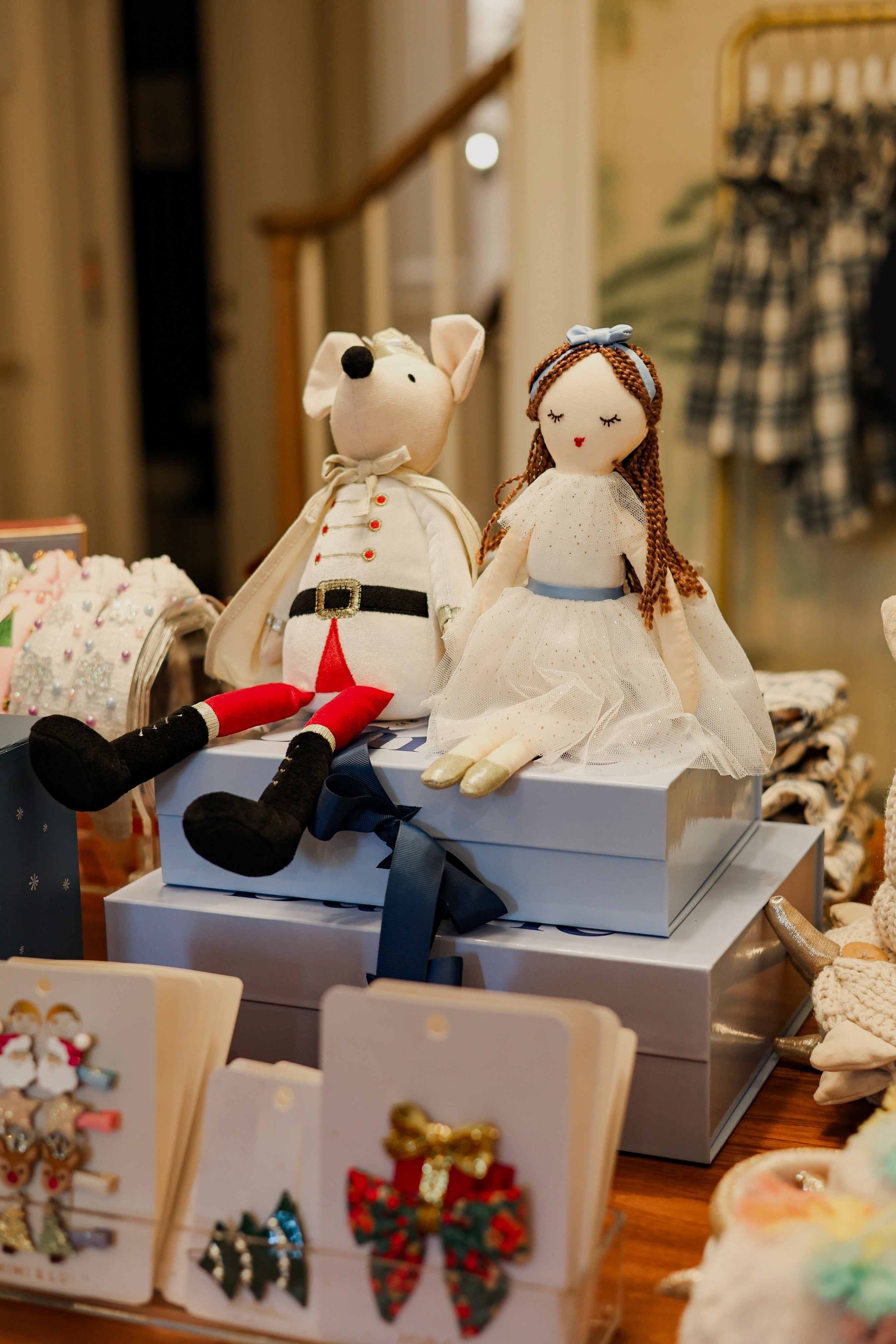 Festive holiday gift display featuring a plush mouse in a cream outfit and a decorative rag doll in a white dress, seated on stacked blue gift boxes with navy ribbons and surrounded by children's toy