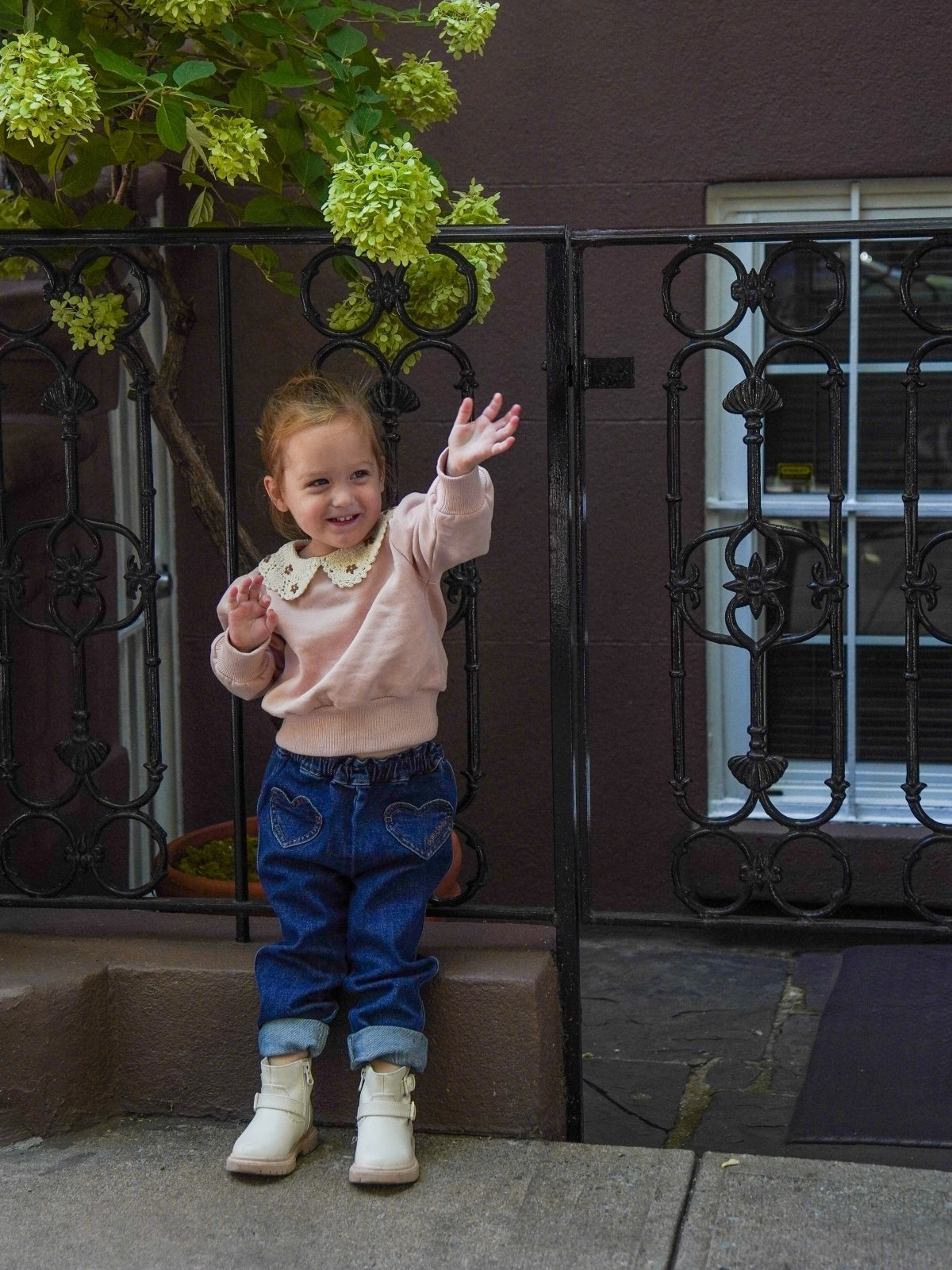 Child standing on a stoop with a decorative iron gate and green flowers in the background