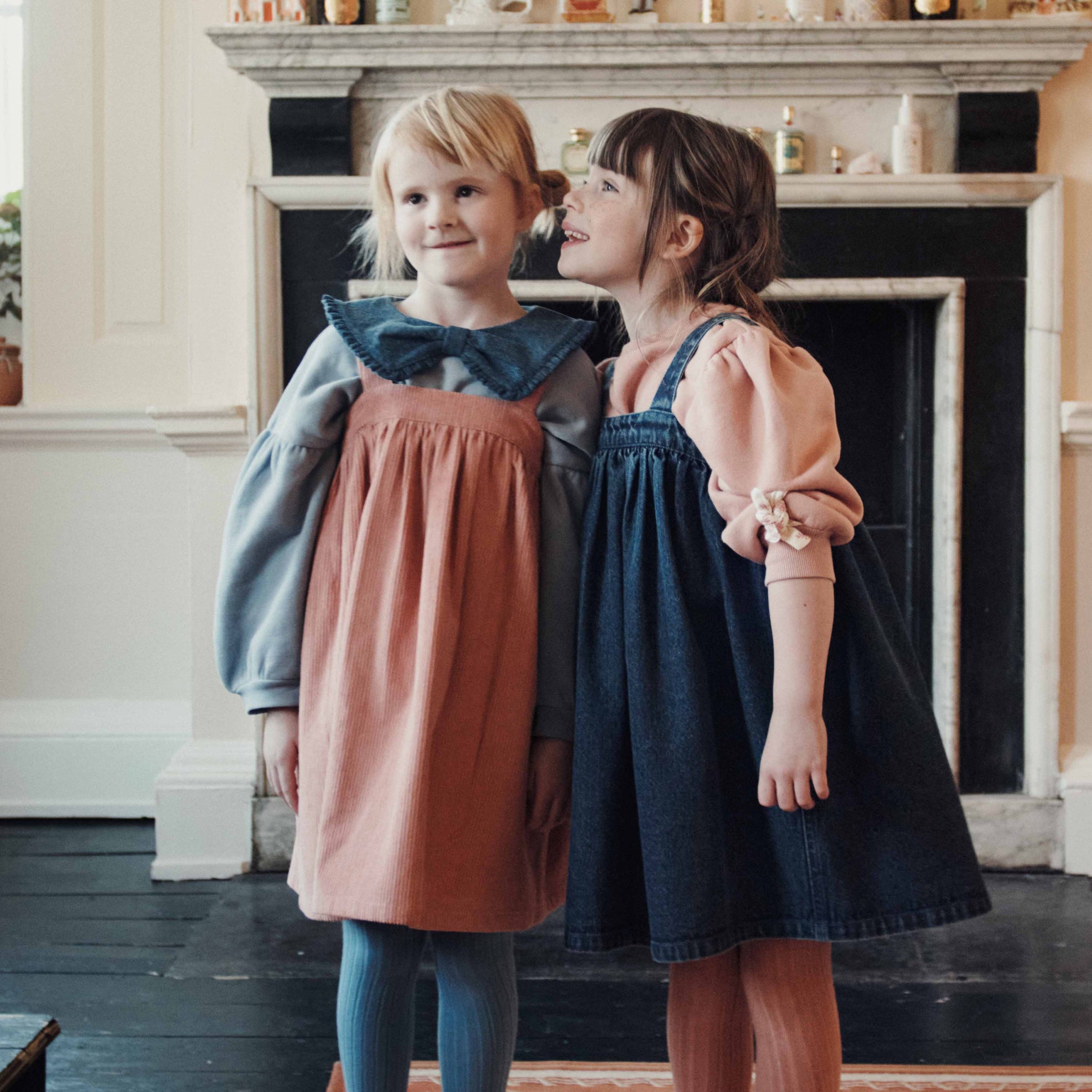 2 girls standing in front of a fireplace, with one girl whispering in her ear