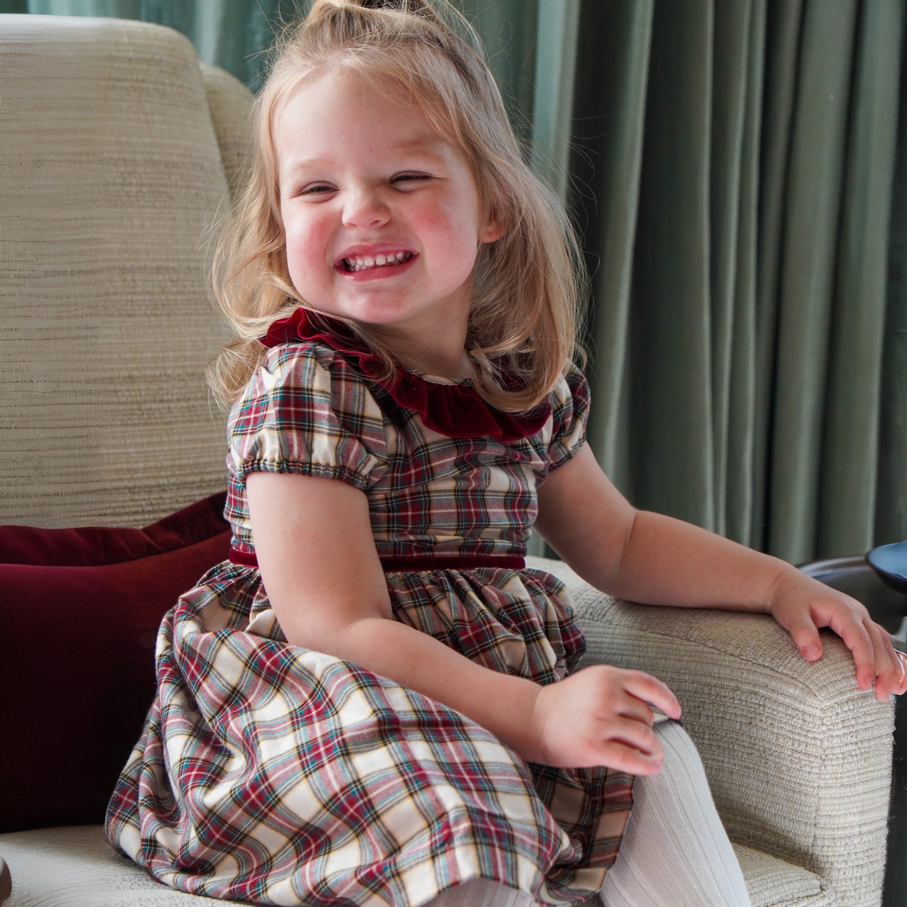 Smiling young girl sitting on a sofa wearing a festive plaid dress with short puff sleeves and a burgundy velvet collar.