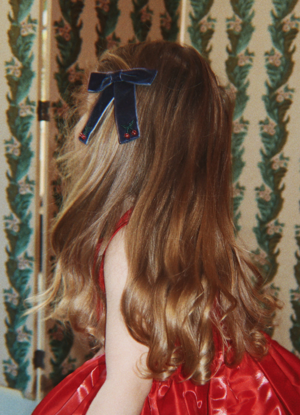 Back view of a young girl with long blonde hair wearing a navy velvet hair bow and a red satin dress.