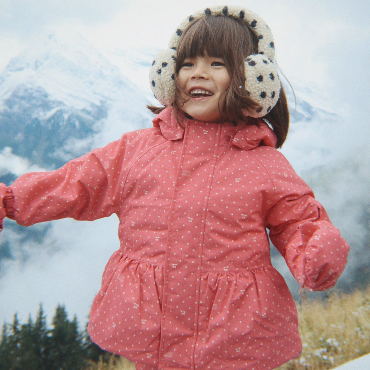 Smiling young girl wearing a pink patterned winter coat with a gathered waist and cream fuzzy polka dot earmuffs, standing outdoors against a snowy mountain backdrop.