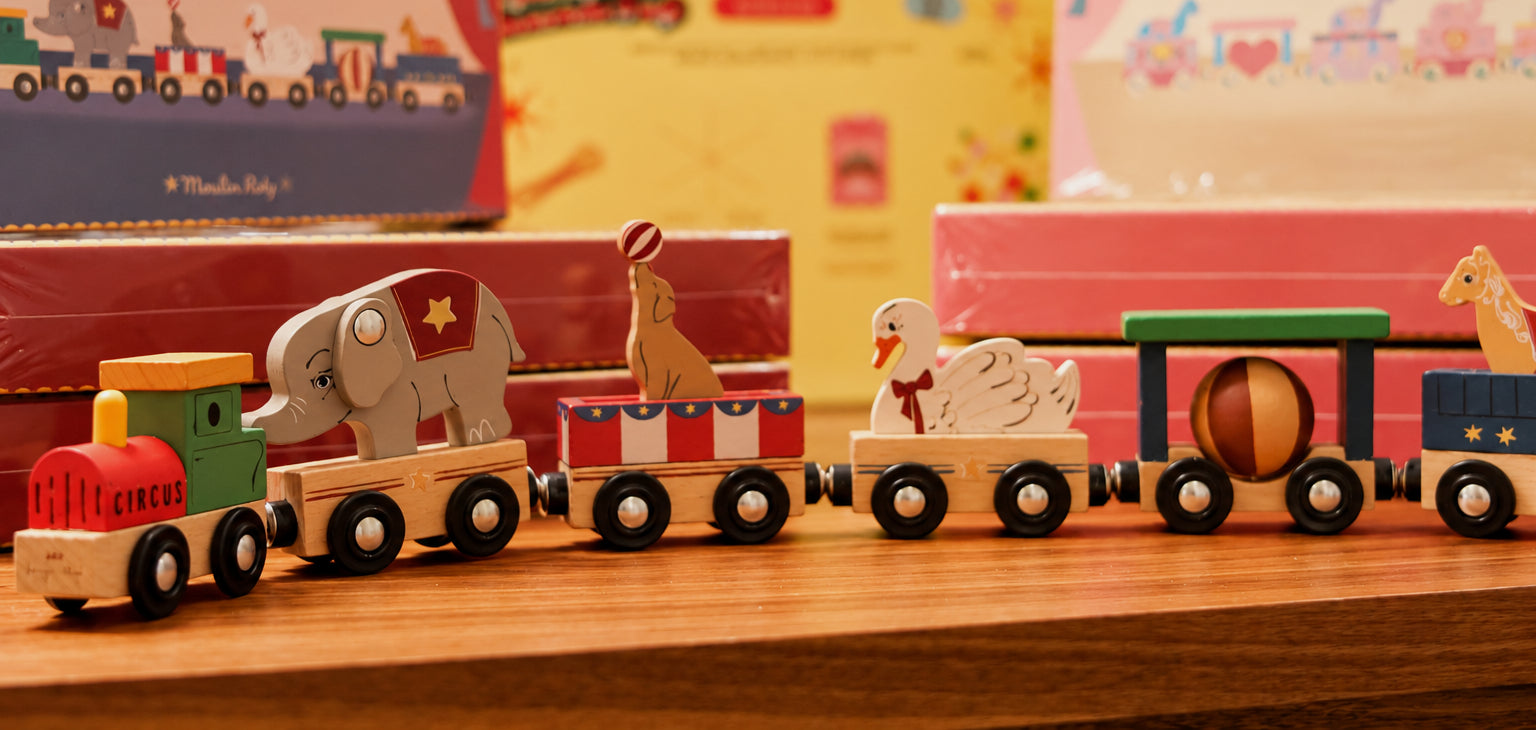 Colorful wooden circus train featuring an elephant, seal, swan, and striped ball, surrounded by festive toy boxes in warm pink, red, and yellow tones.