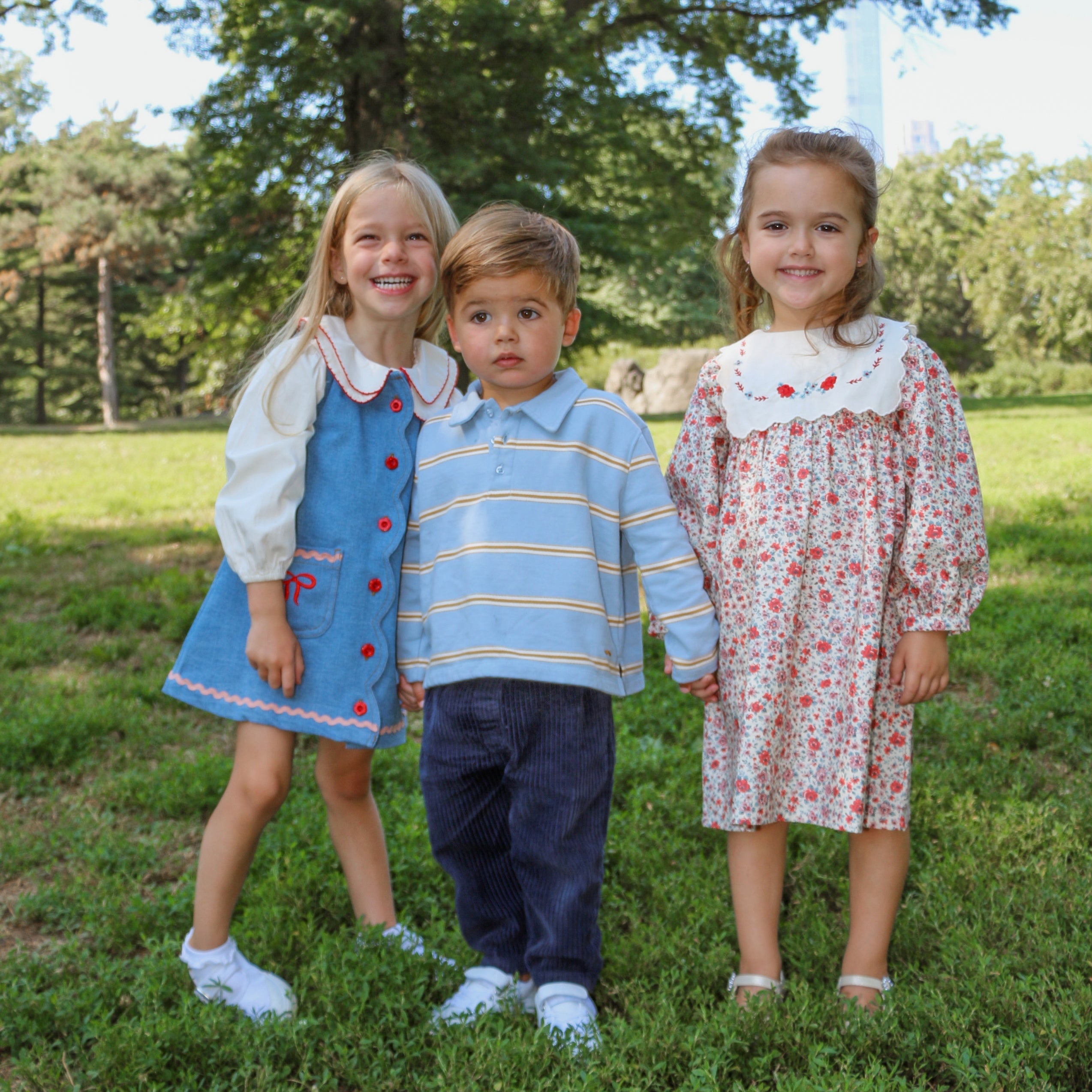 Three children standing together in a grassy area with trees in the background